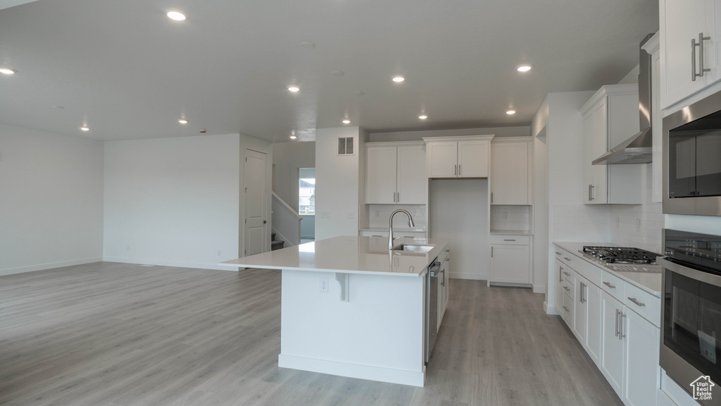 Kitchen with stainless steel appliances, a sink, light wood finished floors, wall chimney range hood, and an island with sink