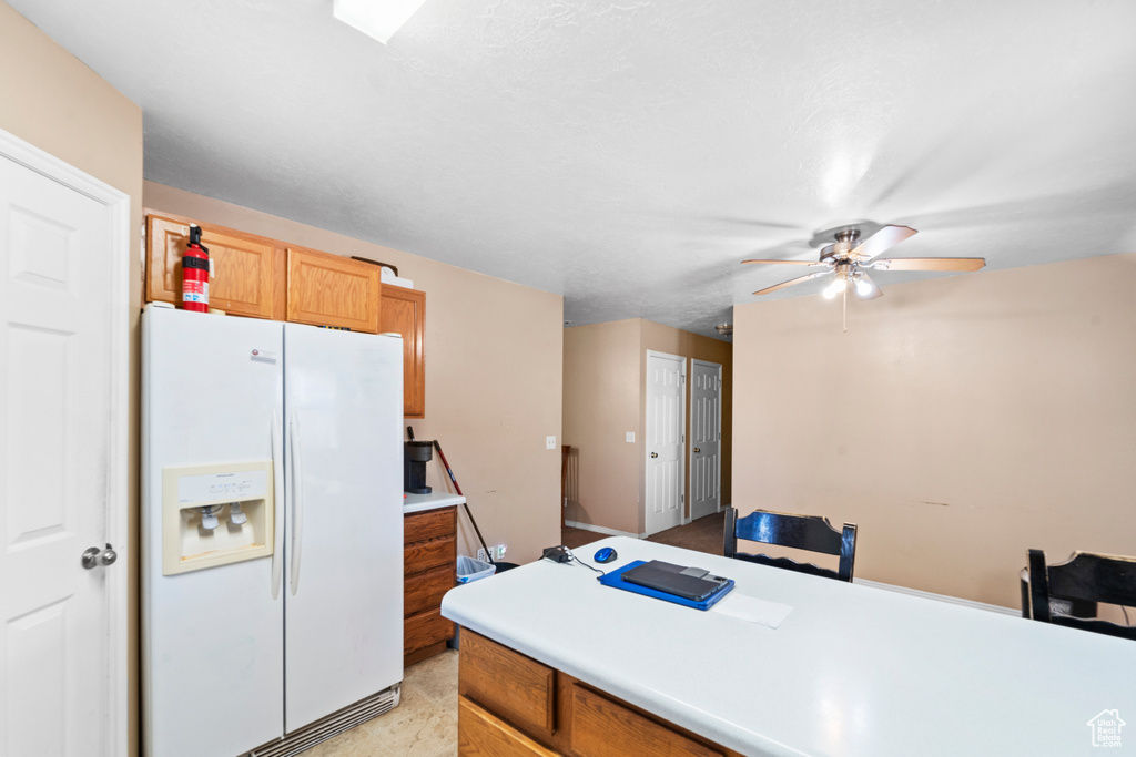 Kitchen featuring white refrigerator with ice dispenser, a ceiling fan, light countertops, and brown cabinetry