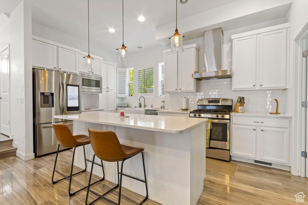 Kitchen with wall chimney range hood, appliances with stainless steel finishes, light wood-type flooring, a kitchen bar, and a kitchen island