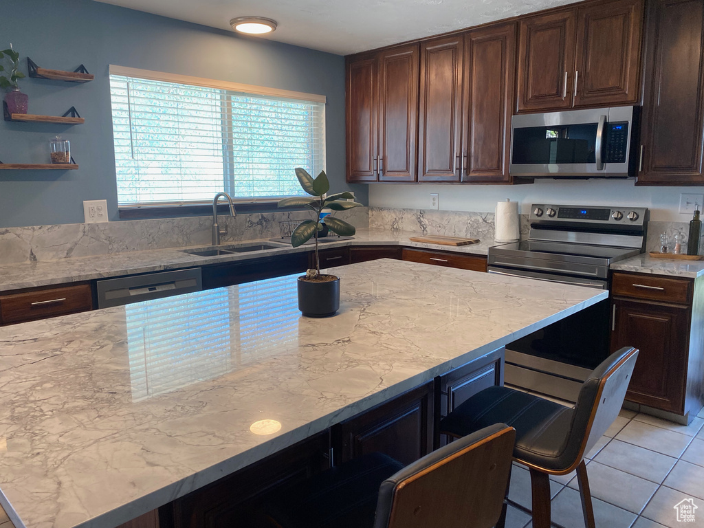 Kitchen with appliances with stainless steel finishes, a sink, a kitchen breakfast bar, light stone counters, and dark brown cabinetry