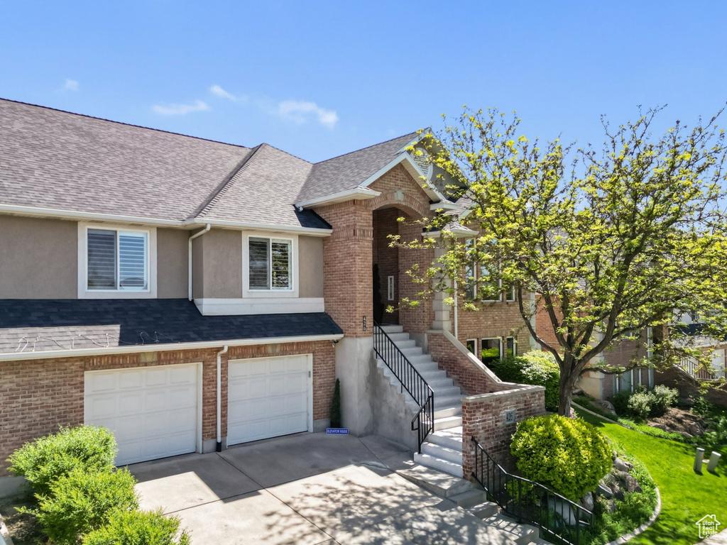 View of property featuring driveway, a garage, roof with shingles, stucco siding, and brick siding