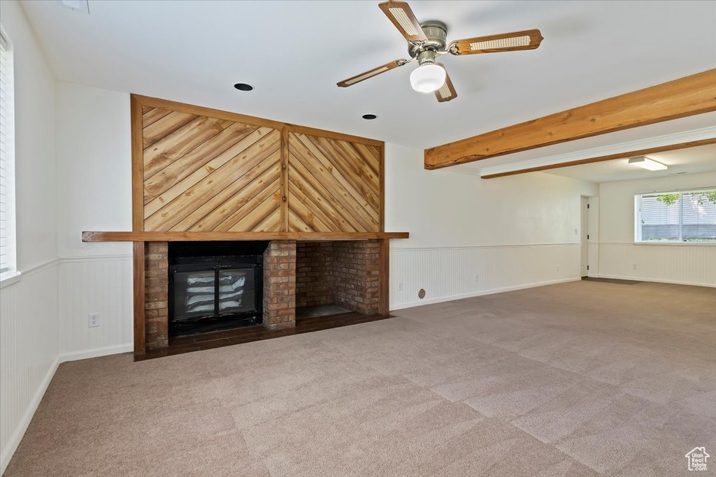 Unfurnished living room featuring carpet, a brick fireplace, ceiling fan, a wainscoted wall, and beamed ceiling