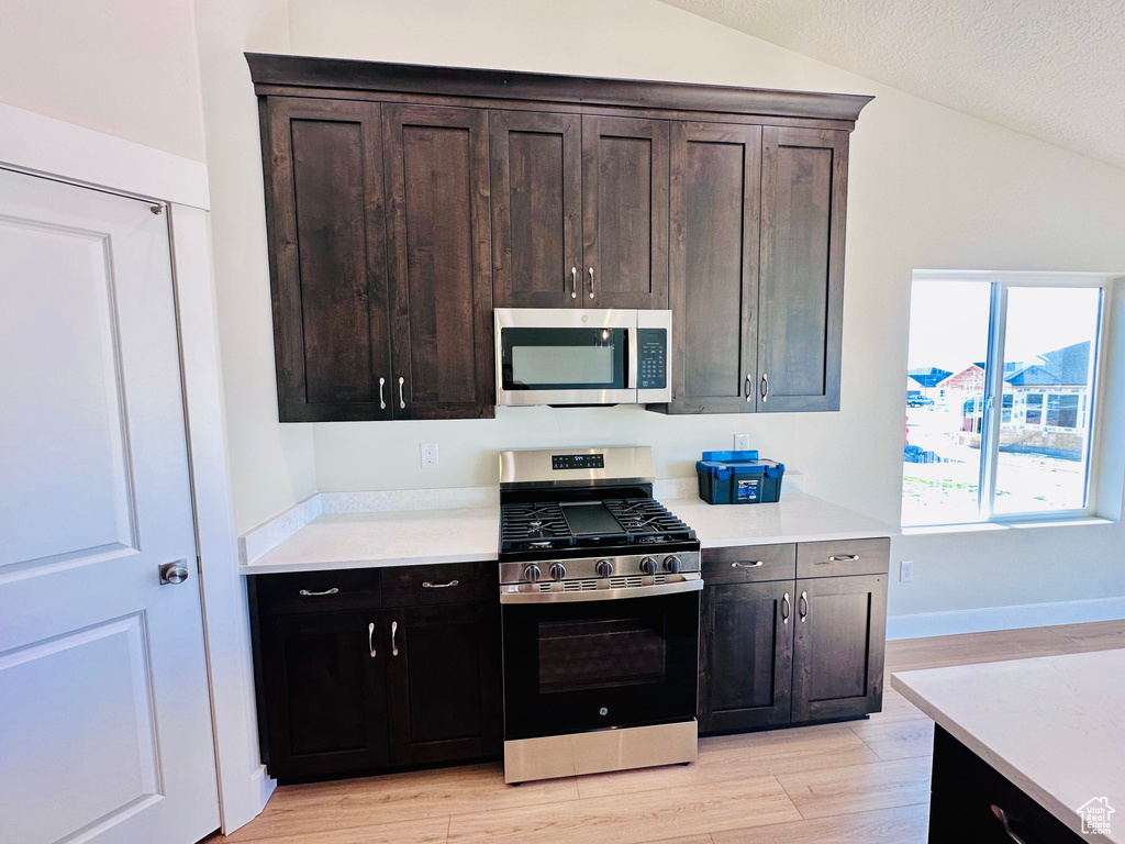 Kitchen with appliances with stainless steel finishes, vaulted ceiling, light wood finished floors, light countertops, and dark brown cabinets