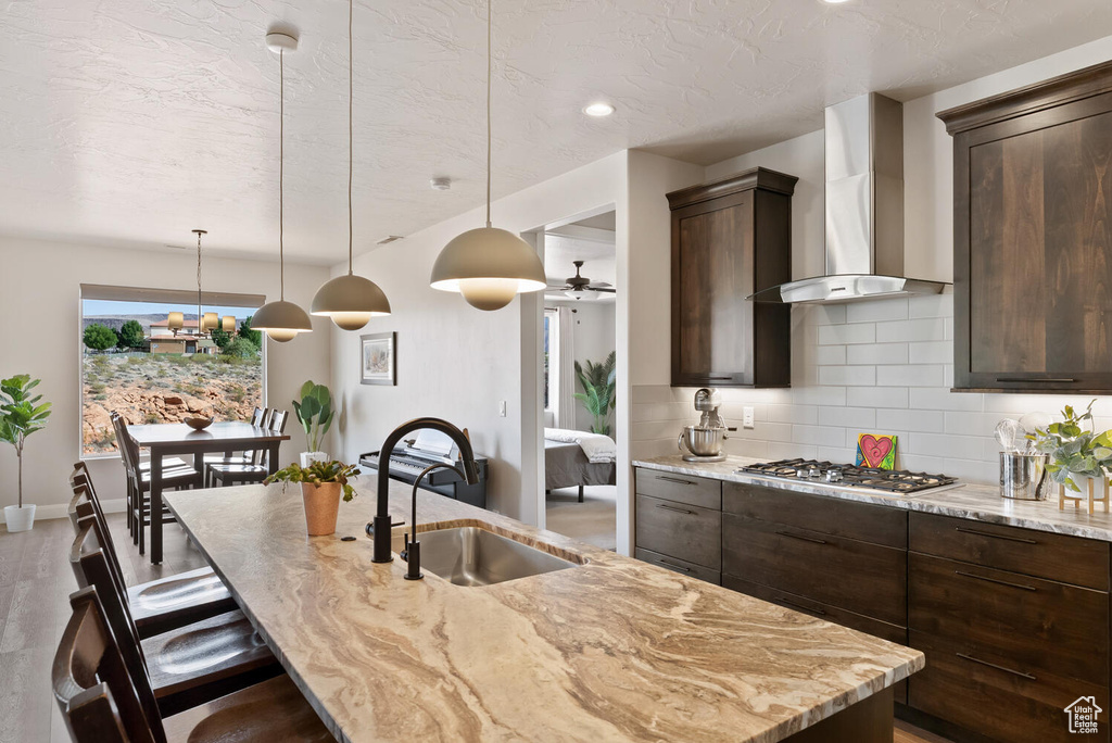 Kitchen with a sink, wall chimney range hood, stainless steel gas cooktop, dark brown cabinetry, and backsplash