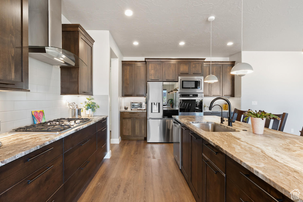 Kitchen with a sink, wall chimney range hood, stainless steel appliances, dark wood-style floors, and light stone counters