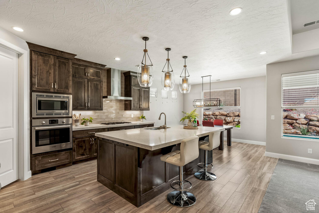 Kitchen featuring stainless steel appliances, wall chimney range hood, a sink, light wood-style floors, and light countertops