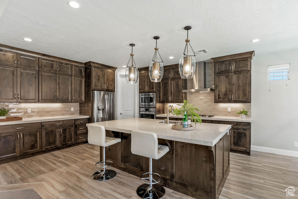 Kitchen featuring wall chimney exhaust hood, appliances with stainless steel finishes, decorative backsplash, light wood-type flooring, and recessed lighting