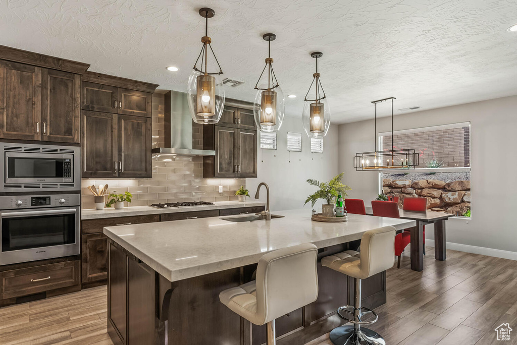 Kitchen with stainless steel appliances, a sink, wall chimney exhaust hood, light wood-style flooring, and decorative backsplash