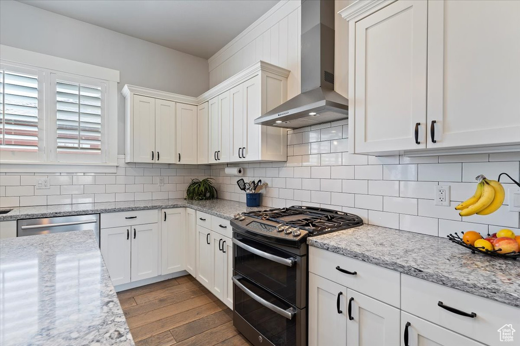 Kitchen featuring stainless steel appliances, wall chimney range hood, hardwood / wood-style flooring, white cabinets, and decorative backsplash