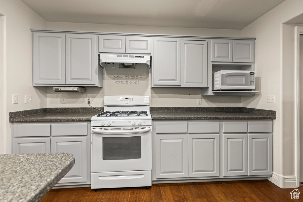 Kitchen featuring white appliances, under cabinet range hood, dark countertops, dark wood-style flooring, and gray cabinetry