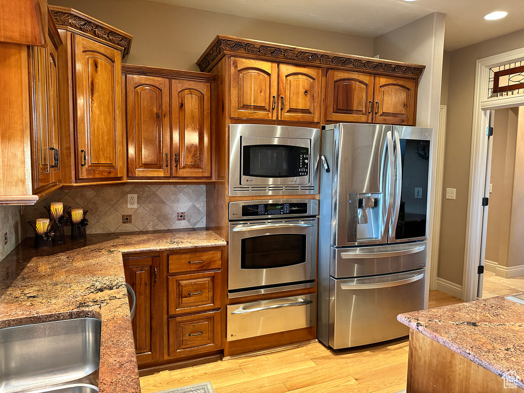 Kitchen featuring stainless steel appliances, a warming drawer, light stone countertops, decorative backsplash, and brown cabinetry