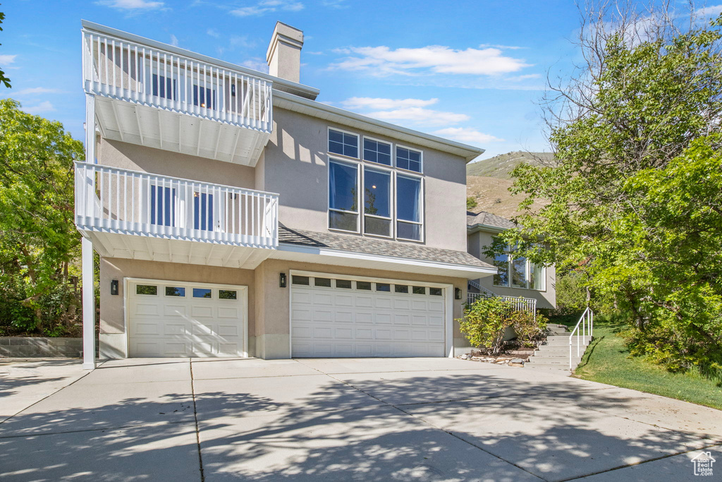 View of front facade featuring a balcony, an attached garage, stucco siding, driveway, and a chimney