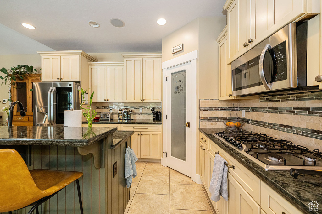 Kitchen with stainless steel appliances, cream cabinets, a kitchen breakfast bar, backsplash, and dark stone counters