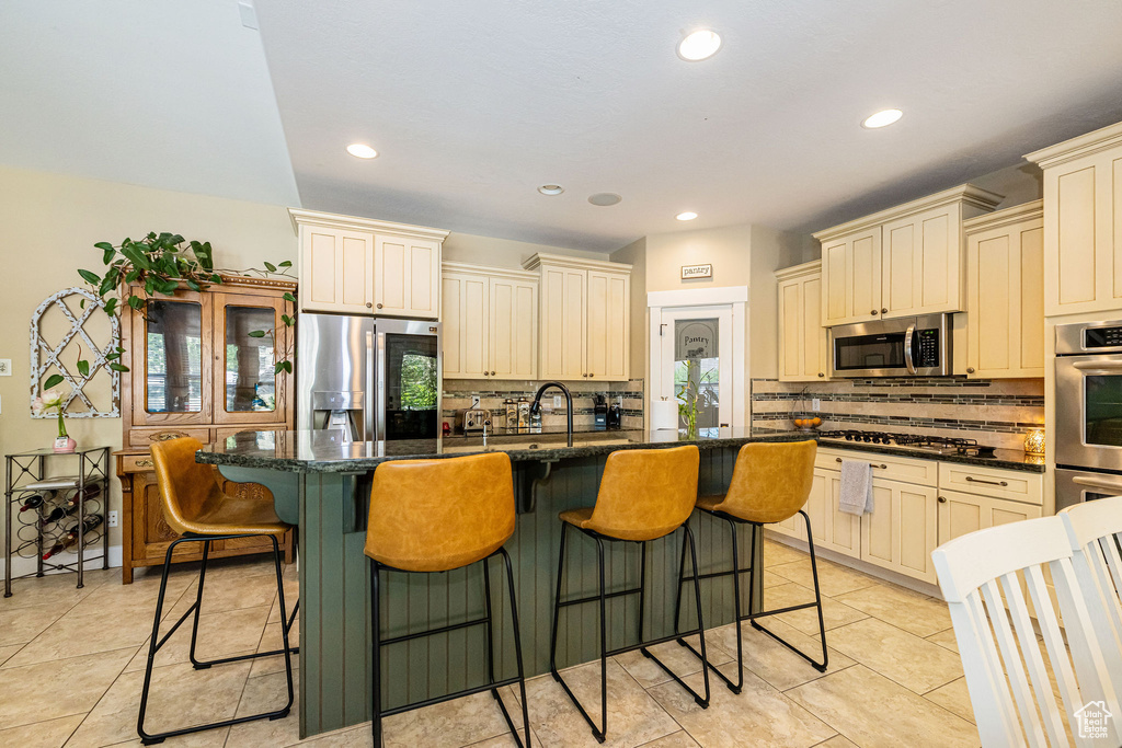 Kitchen with stainless steel appliances, cream cabinetry, tasteful backsplash, light tile patterned floors, and a kitchen island with sink
