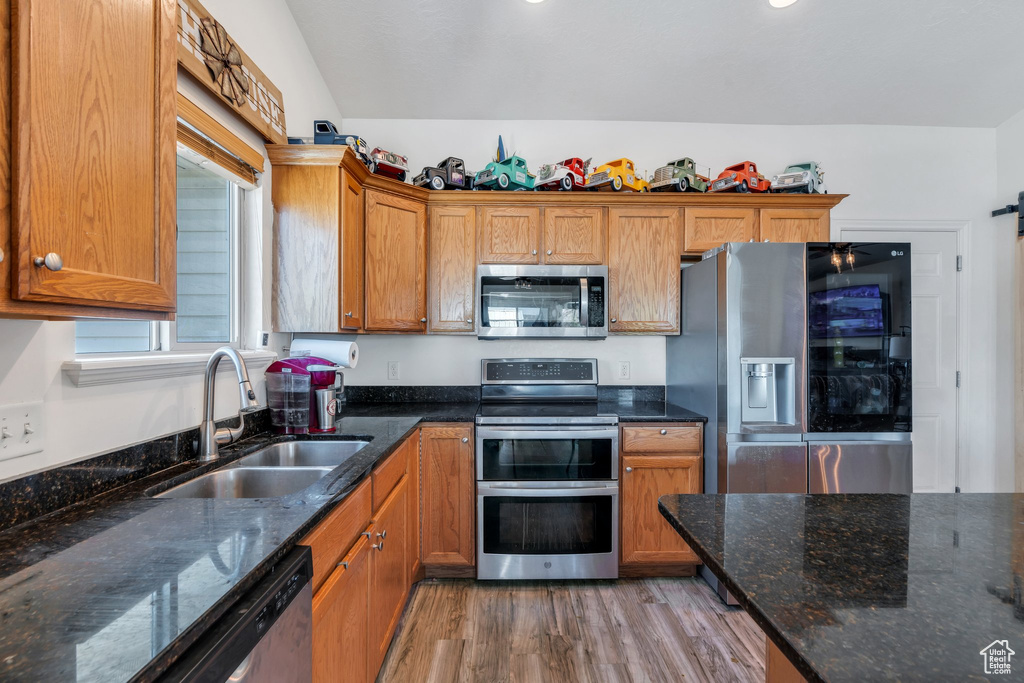 Kitchen with appliances with stainless steel finishes, a sink, light wood-style flooring, brown cabinets, and dark stone countertops