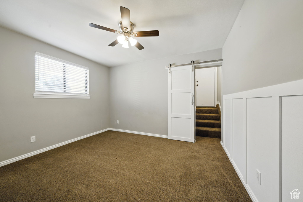 Unfurnished bedroom with a barn door, dark colored carpet, ceiling fan, and baseboards