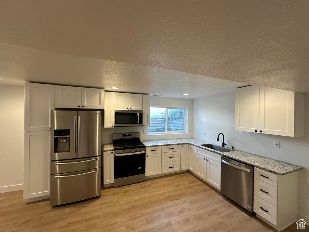Kitchen with stainless steel appliances, a sink, white cabinetry, light stone countertops, and light wood-type flooring