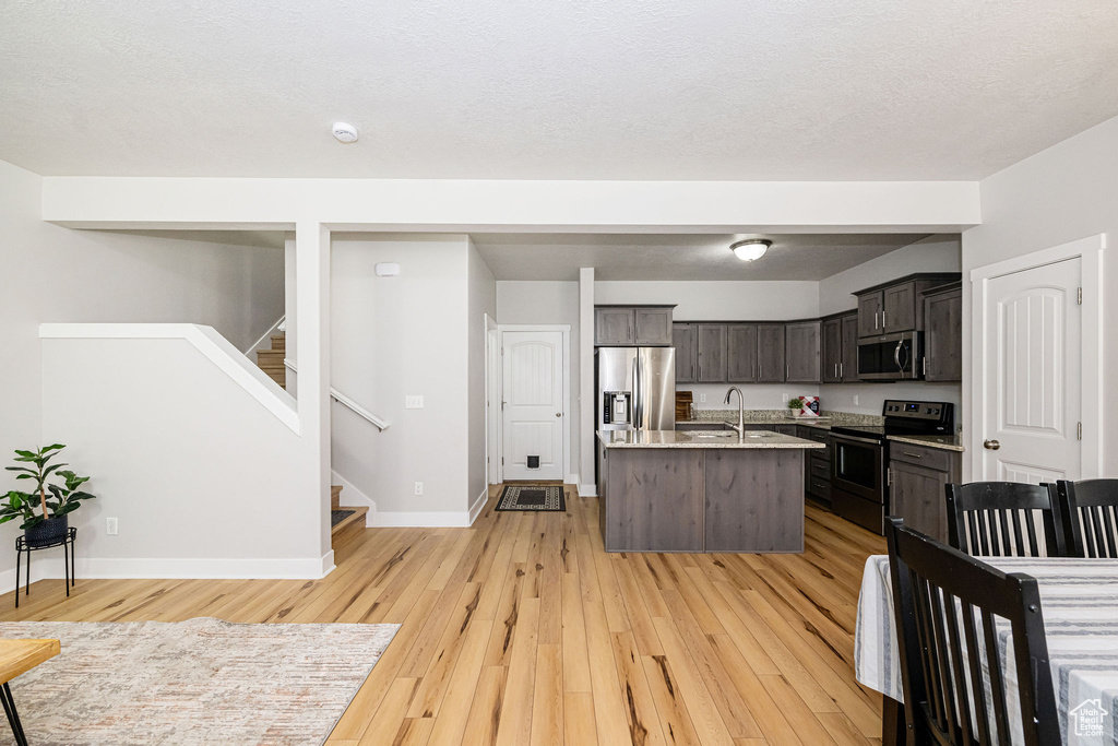 Kitchen featuring stainless steel appliances, light wood-style floors, a center island with sink, baseboards, and dark brown cabinetry