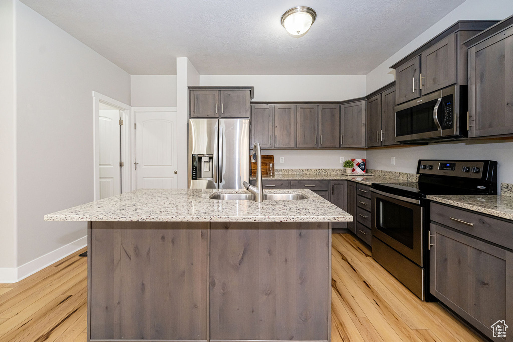 Kitchen featuring stainless steel appliances, a sink, light wood finished floors, dark brown cabinets, and an island with sink