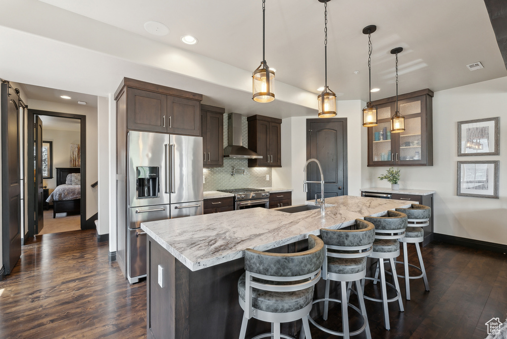Kitchen with high quality appliances, a sink, wall chimney exhaust hood, dark brown cabinetry, and glass insert cabinets