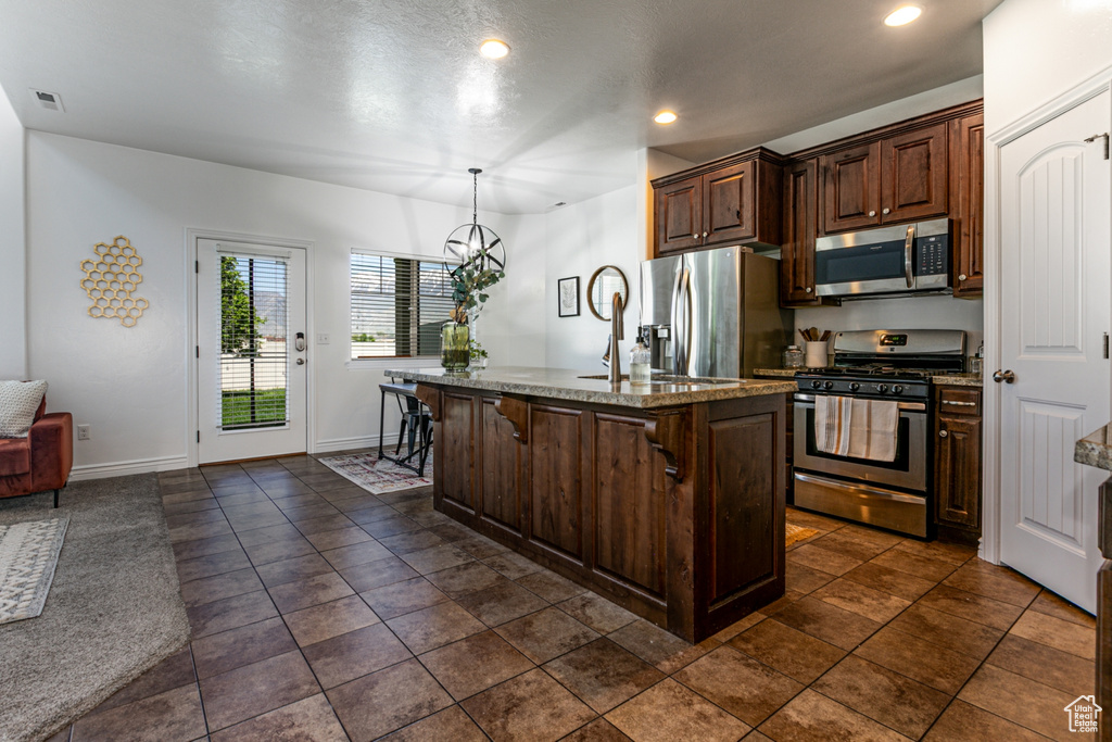 Kitchen featuring appliances with stainless steel finishes, a chandelier, light stone counters, a center island with sink, and decorative light fixtures