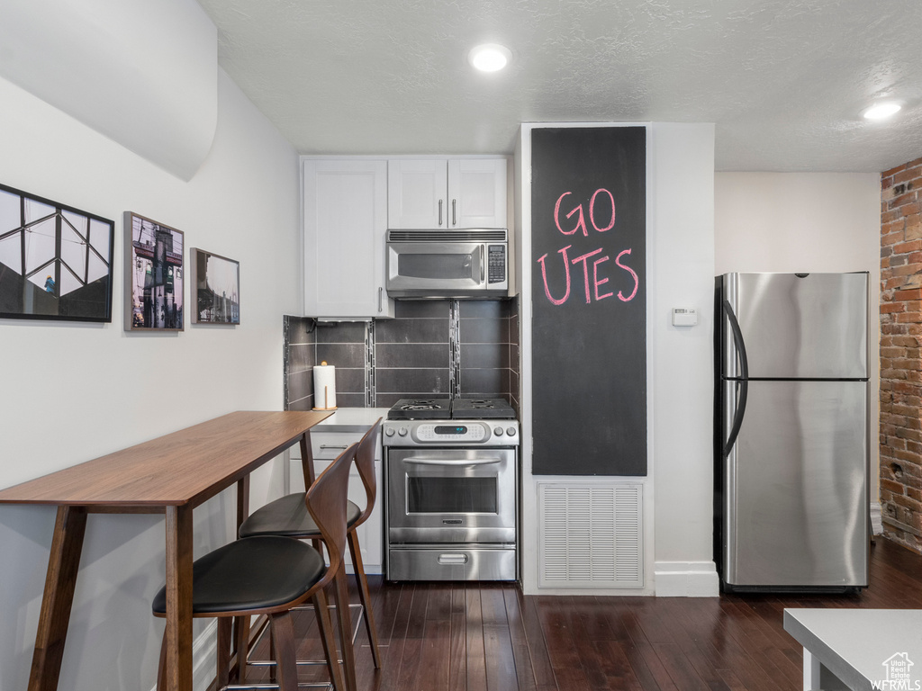 Kitchen with appliances with stainless steel finishes, white cabinets, decorative backsplash, brick wall, and dark wood-type flooring