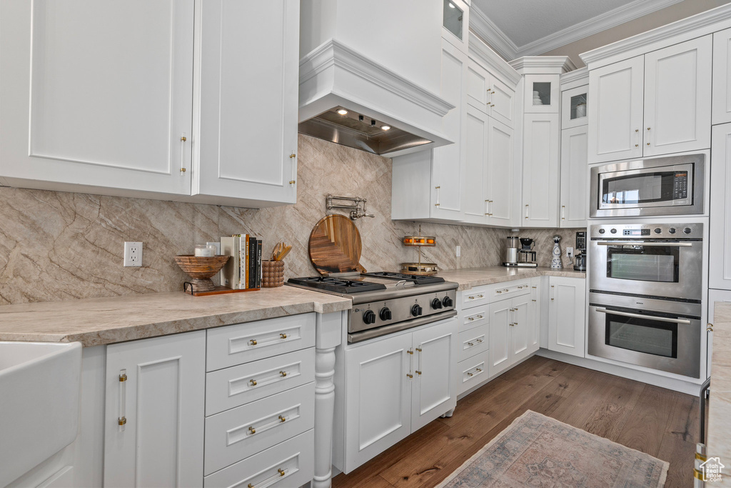 Kitchen with stainless steel appliances, custom range hood, ornamental molding, white cabinetry, and dark wood finished floors