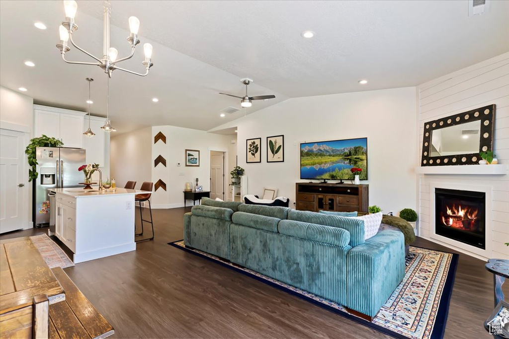 Living room featuring dark wood-style flooring, lofted ceiling, ceiling fan, a large fireplace, and a chandelier
