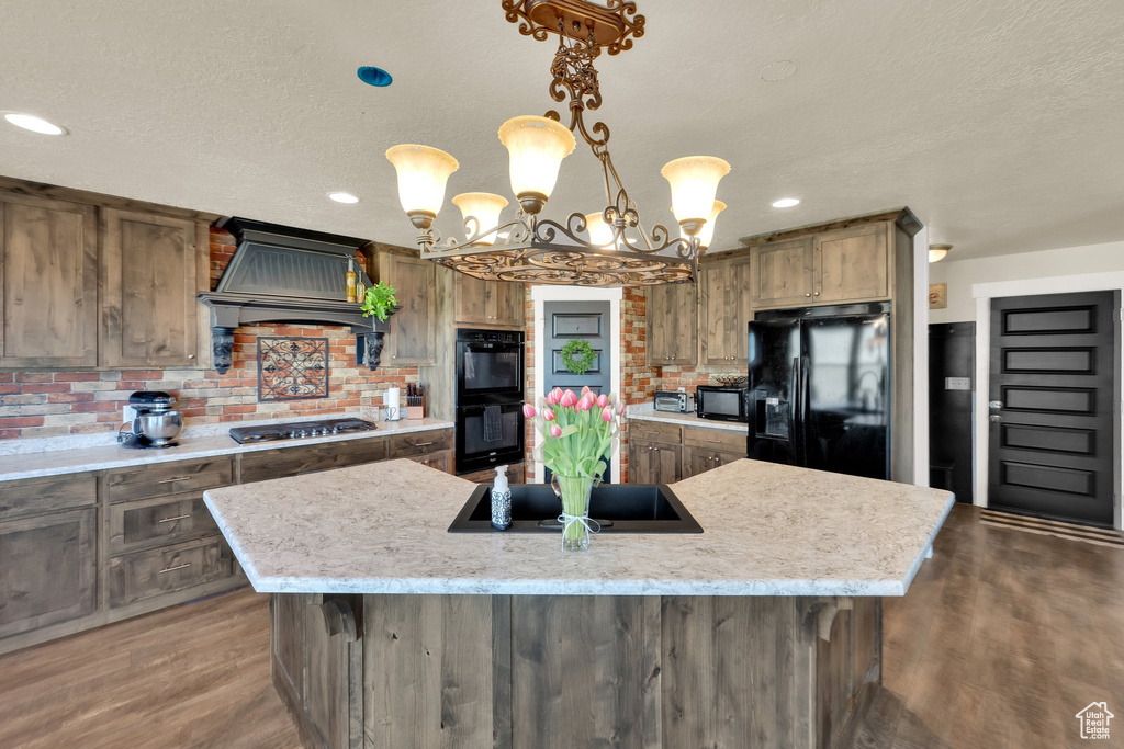 Kitchen with black appliances, dark wood finished floors, custom range hood, an island with sink, and a chandelier