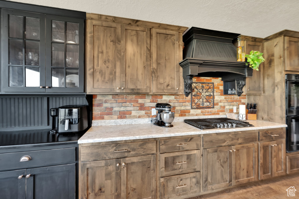 Kitchen featuring stainless steel gas cooktop, custom range hood, glass insert cabinets, light stone countertops, and a textured ceiling