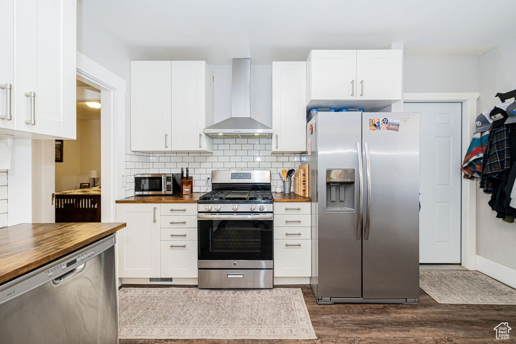 Kitchen with appliances with stainless steel finishes, butcher block counters, wall chimney exhaust hood, dark wood-type flooring, and tasteful backsplash