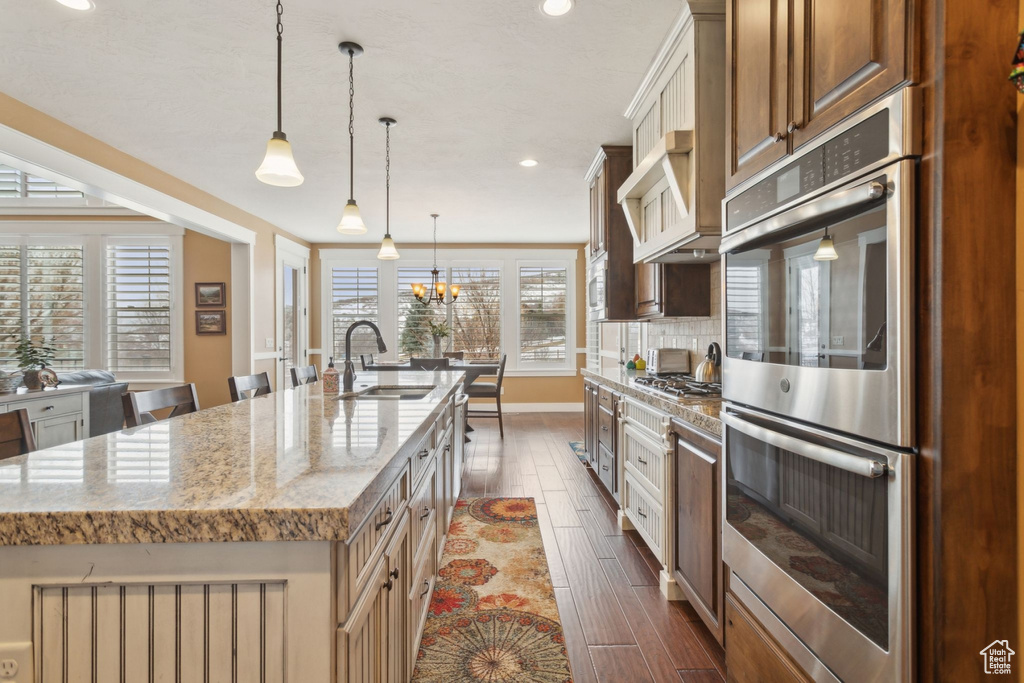 Kitchen featuring appliances with stainless steel finishes, a sink, dark wood-style flooring, a kitchen island with sink, and light stone countertops