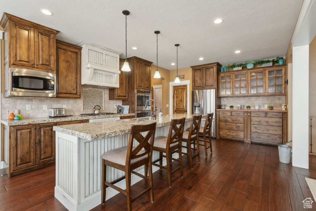 Kitchen featuring stainless steel appliances, brown cabinetry, tasteful backsplash, a kitchen island with sink, and dark wood-style floors