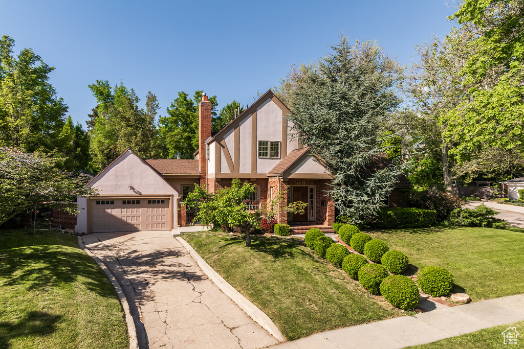 Tudor-style house featuring stucco siding, a front lawn, a chimney, driveway, and a garage