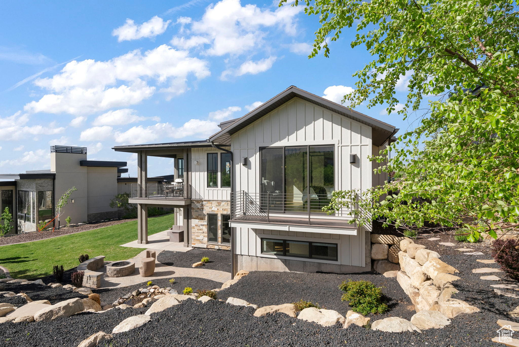 Rear view of property featuring board and batten siding, an outdoor fire pit, stone siding, a patio, and a balcony