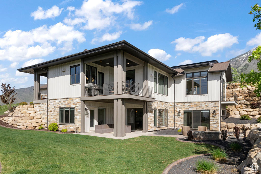 Back of property featuring a balcony, stone siding, a patio, a lawn, and a mountain view