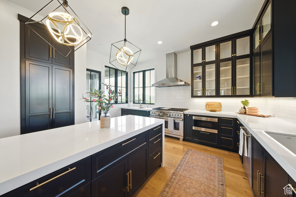 Kitchen featuring wall chimney exhaust hood, appliances with stainless steel finishes, light countertops, decorative backsplash, and pendant lighting