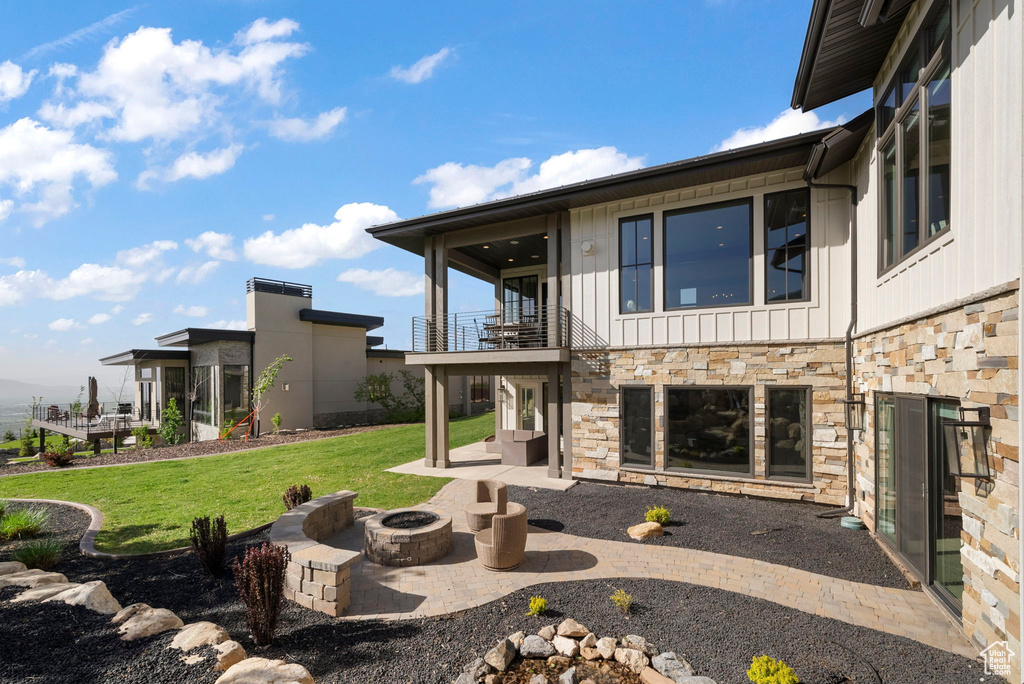 Back of house featuring a balcony, stone siding, an outdoor fire pit, a lawn, and a patio