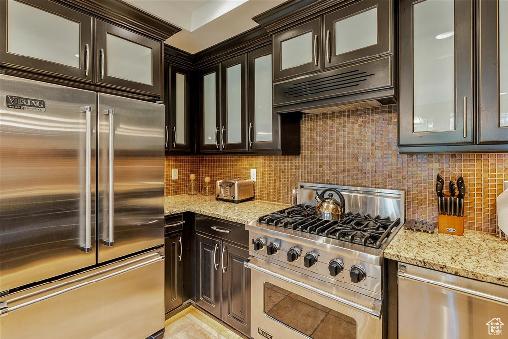 Kitchen with premium appliances, decorative backsplash, light stone counters, and dark brown cabinets