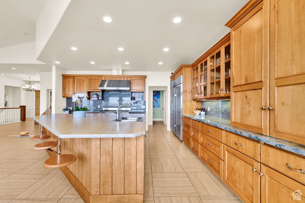 Kitchen featuring stainless steel built in fridge, decorative backsplash, wall chimney range hood, a kitchen island with sink, and a sink
