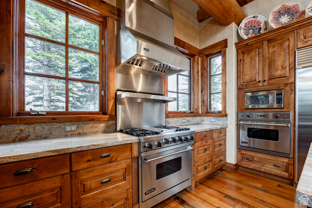 Kitchen featuring built in appliances, light wood-type flooring, light stone countertops, exhaust hood, and brown cabinets