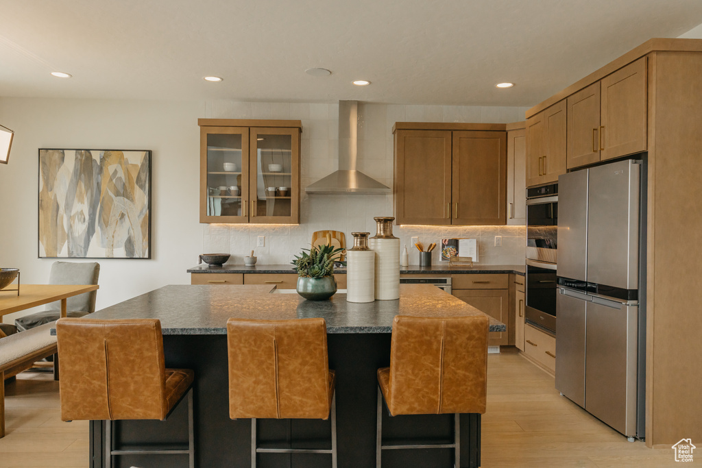 Kitchen featuring stainless steel appliances, wall chimney range hood, dark countertops, a kitchen island, and light wood-type flooring