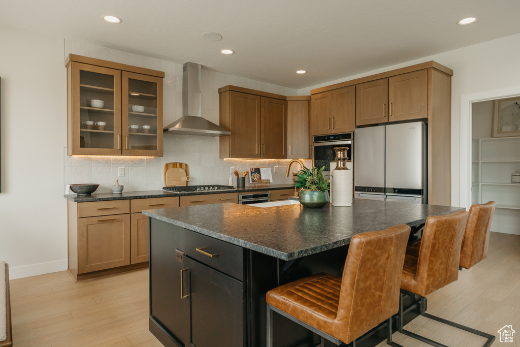 Kitchen featuring wall chimney range hood, appliances with stainless steel finishes, light wood-style flooring, tasteful backsplash, and glass insert cabinets