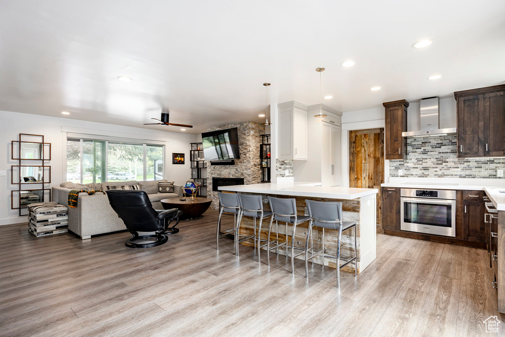 Kitchen featuring stainless steel oven, wall chimney range hood, open floor plan, dark brown cabinetry, and light wood-style floors