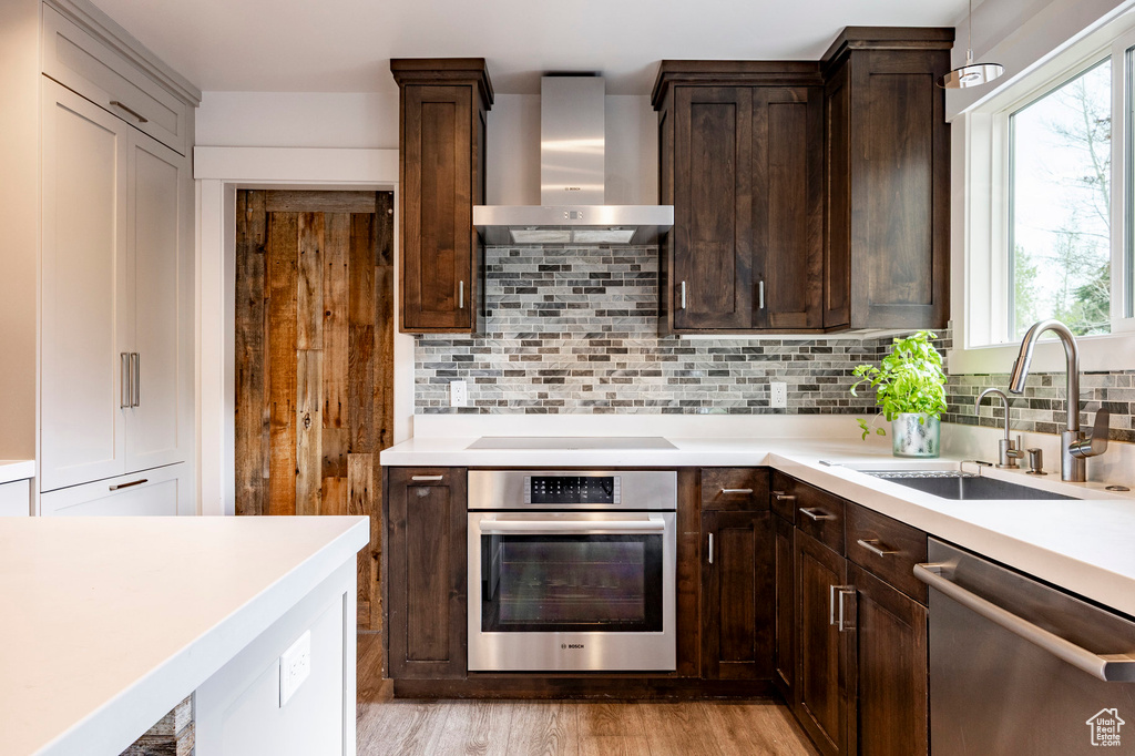 Kitchen with appliances with stainless steel finishes, wall chimney range hood, a sink, dark brown cabinets, and light countertops