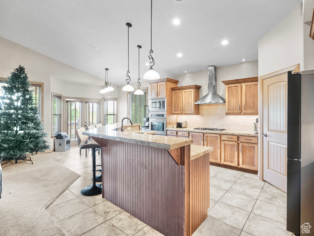 Kitchen featuring wall chimney range hood, appliances with stainless steel finishes, tasteful backsplash, a kitchen island with sink, and light tile patterned flooring