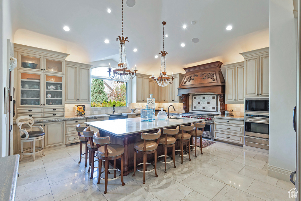 Kitchen with appliances with stainless steel finishes, custom range hood, cream cabinetry, an island with sink, and vaulted ceiling