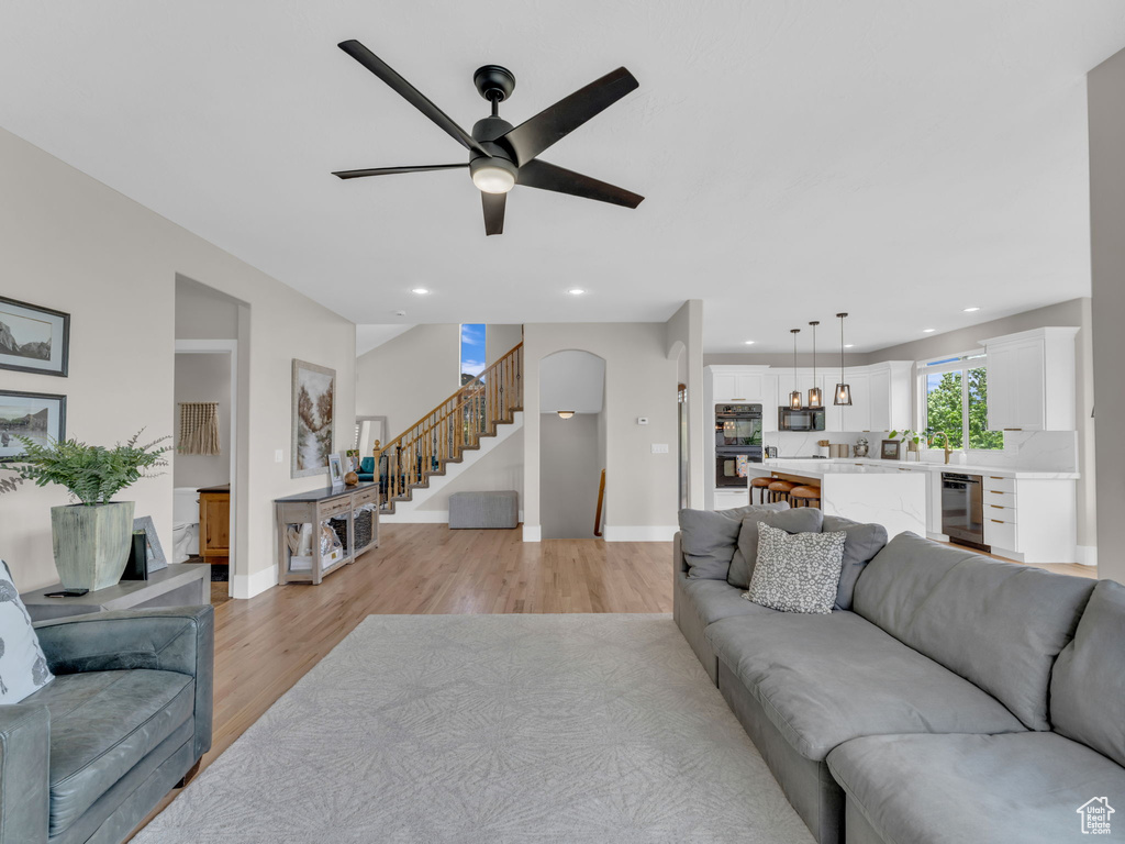 Living room with beverage cooler, light wood-style floors, a ceiling fan, baseboards, and stairway