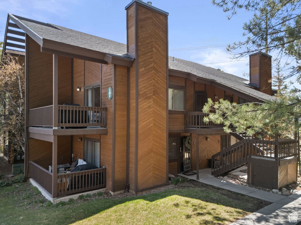 Rear view of property featuring a chimney, a balcony, a shingled roof, and a lawn