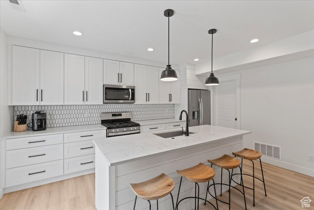 Kitchen with stainless steel appliances, a sink, a center island with sink, backsplash, and light wood-style flooring